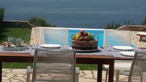 Een eettafel met uitzicht op zee, een fruitmand en een placemat met een kustlandschap.