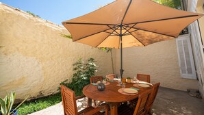 A round wooden table with a large beige umbrella, surrounded by wooden chairs, set against a textured wall with a potted plant nearby. - Roma Divine: home cinema, luxury city center (Nimes)