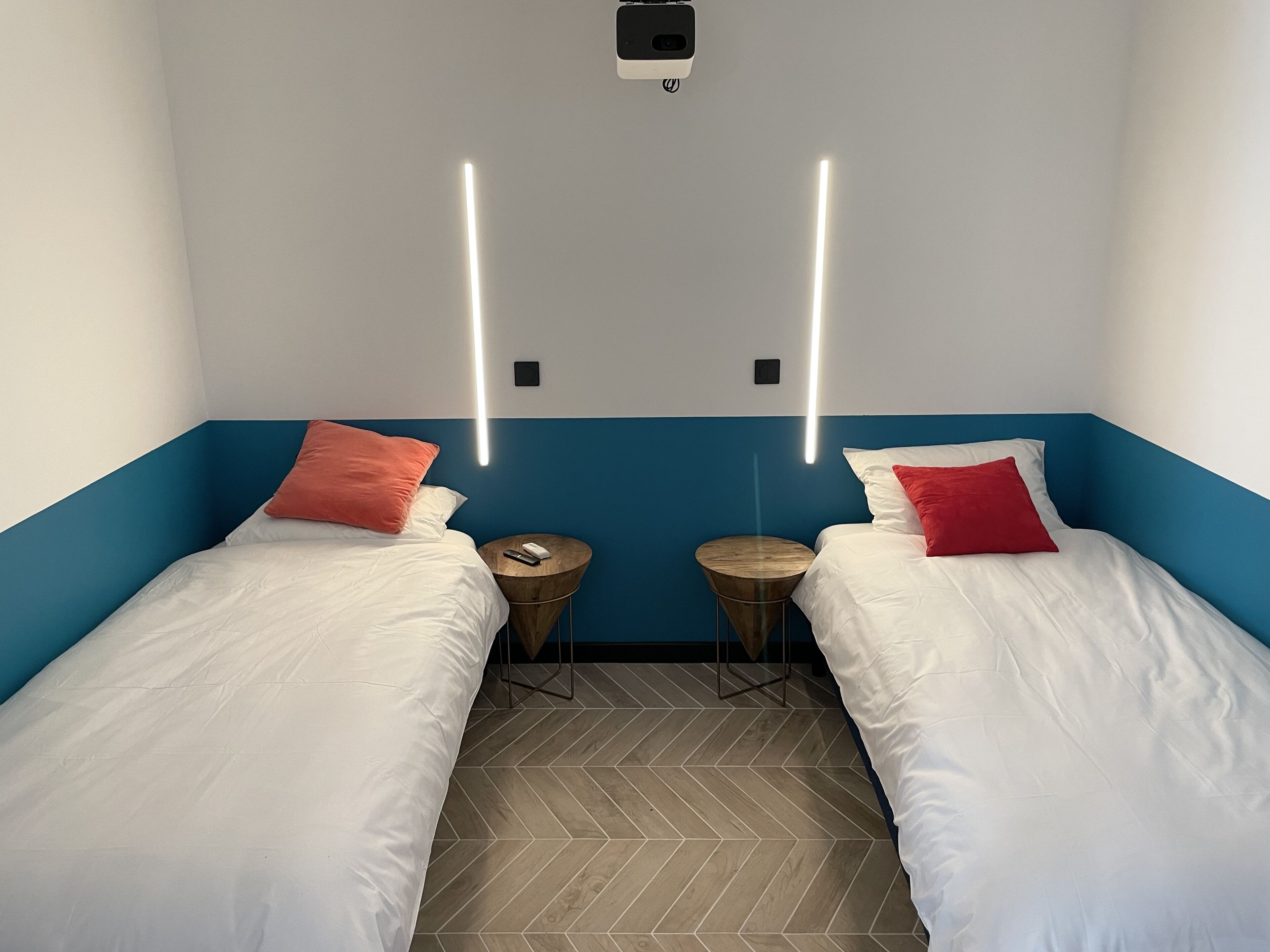A hotel room with two single beds, each with white bedding and a red pillow. There are two wooden nightstands with a phone and a charger on each. The room has a blue accent wall and a herringbone-patterned wooden floor.