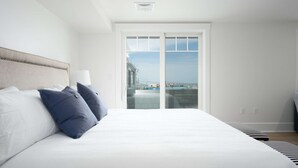 A bedroom with a large bed, a headboard, a nightstand, and a window with a view of a building and sky.