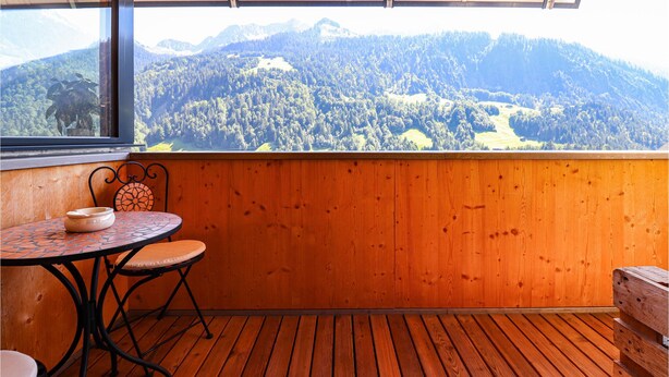 A balcony with a wooden floor and a table with two chairs. A large window offers a view of a forested mountain range. - Ferienwohnung Sonntag (Sonntag)
