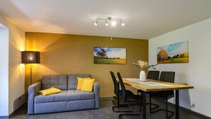 A modern room with a grey sofa, a wooden dining table with chairs, a wall-mounted lamp, and two landscape paintings. - Ferienwohnung Sonntag (Sonntag)