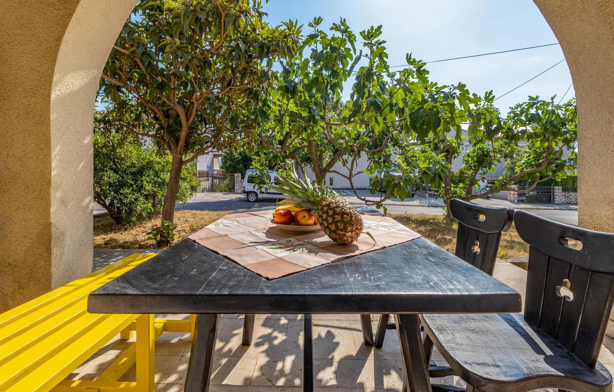 An outdoor table with a pineapple and a plate of fruits, surrounded by trees and benches.