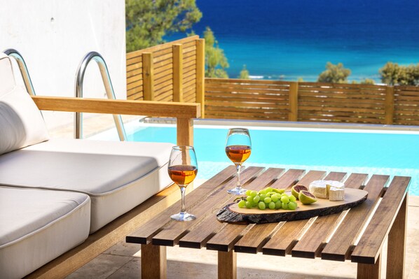Escena junto a la piscina con una mesa de madera, dos copas de vino, una tabla de quesos y un sofá blanco.