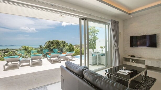 A modern living room with a large glass door leading to a balcony with outdoor furniture and a view of the ocean.