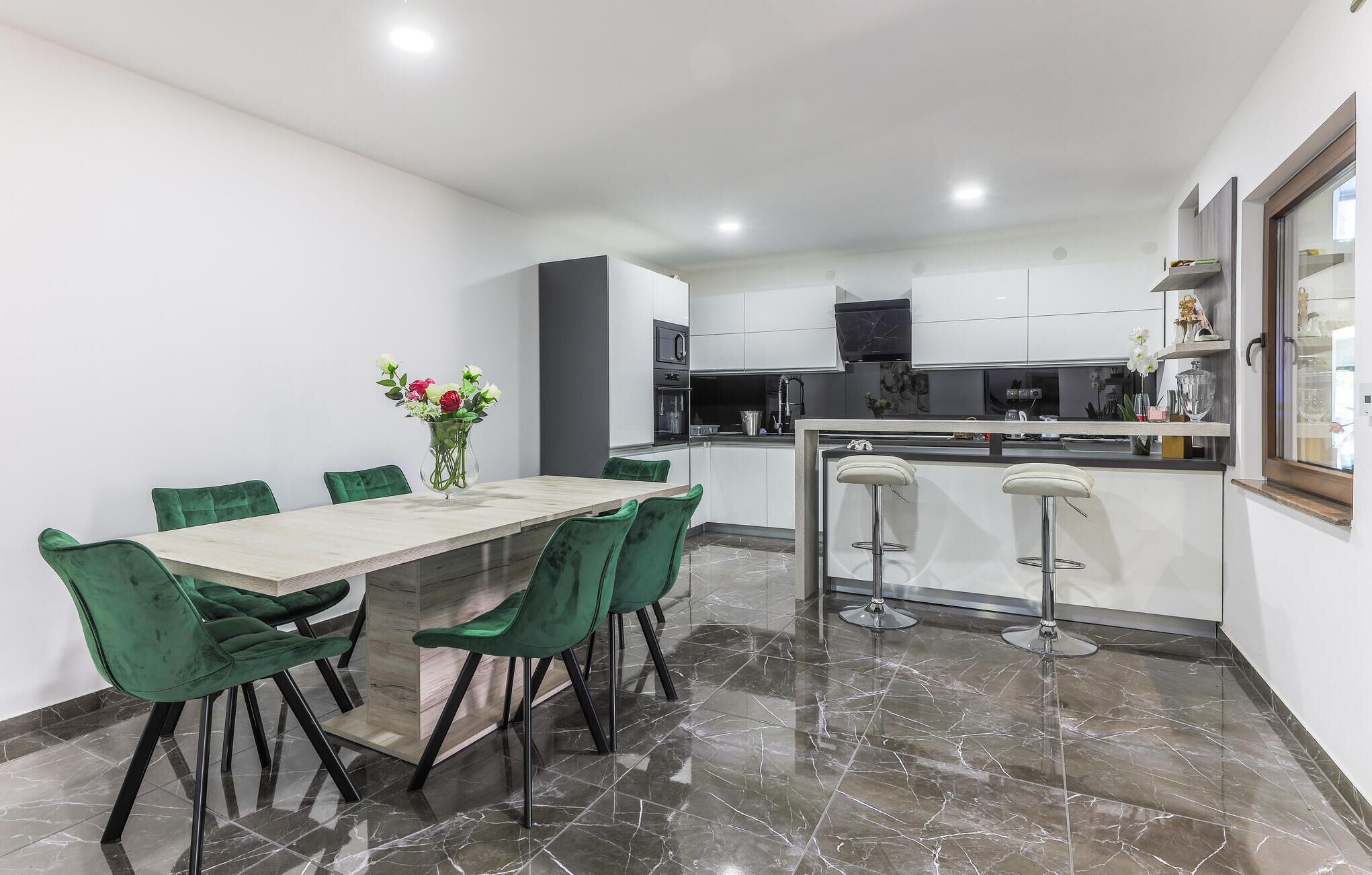 Una cocina moderna con armarios blancos, encimera de mármol y comedor con mesa de madera y sillas verdes.