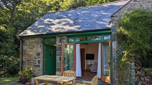 Une petite maison en pierre avec une porte et des fenêtres vertes, une table et un banc en bois à l’extérieur, et un téléviseur dans la pièce.