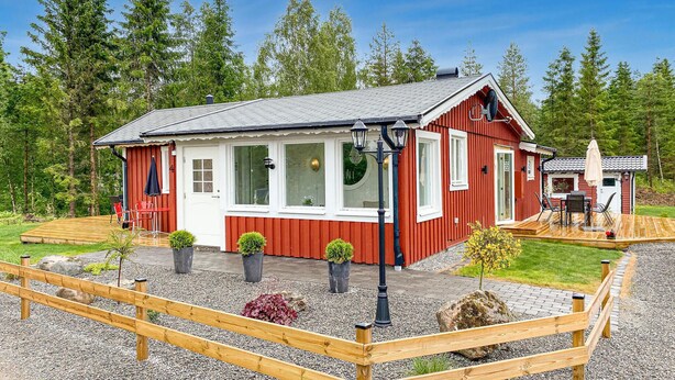 A red and white cabin with a deck, surrounded by a wooden fence and gravel, set in a forested area. - 2 bedroom lovely home in Gislaved (Gislaved)
