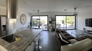 A modern kitchen with a central island, a dining area with a glass table, a sofa, and a TV mounted on the wall.