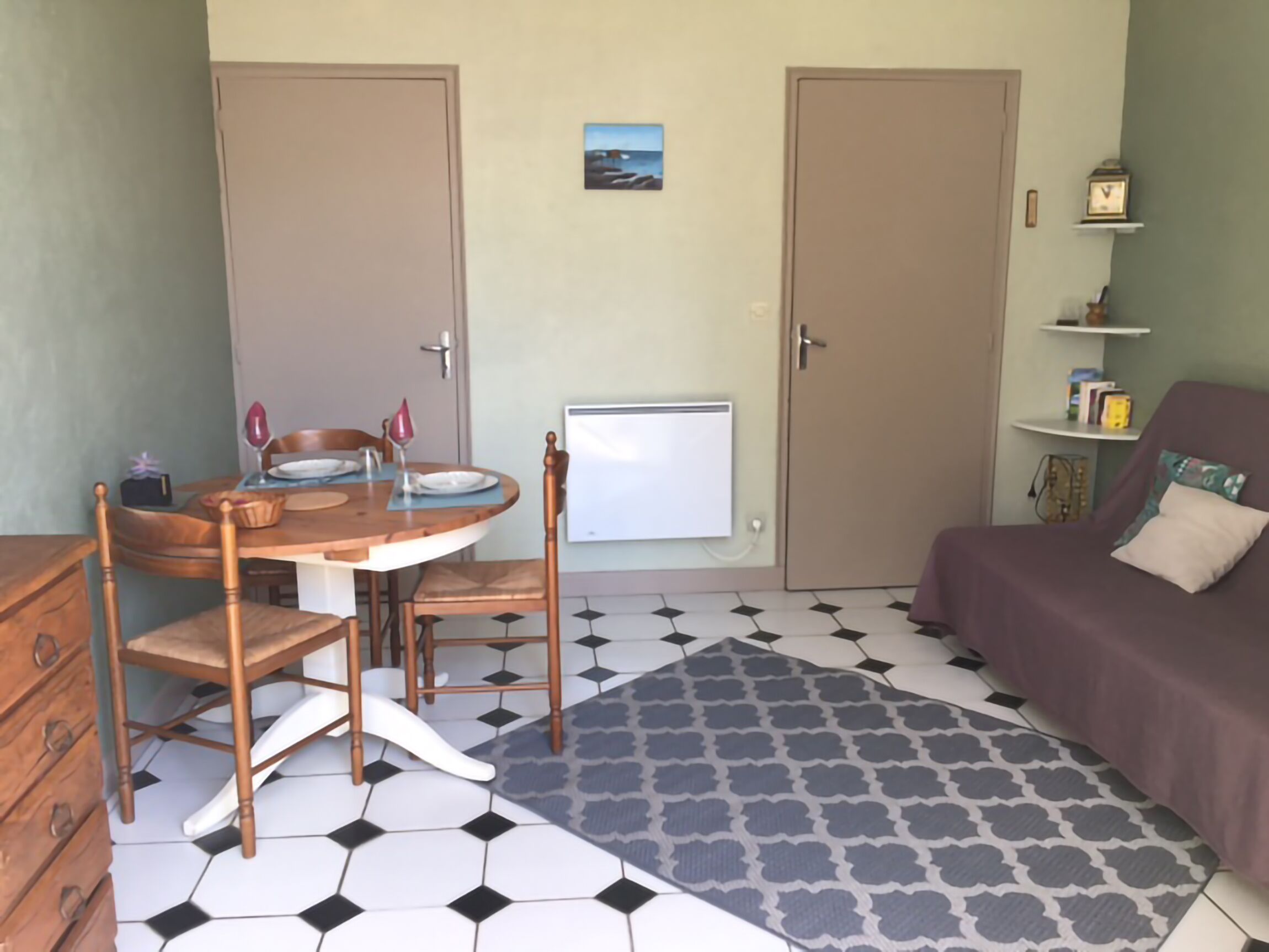 A compact room with a dining table, chairs, a sofa, a patterned rug, and a small shelf with books.