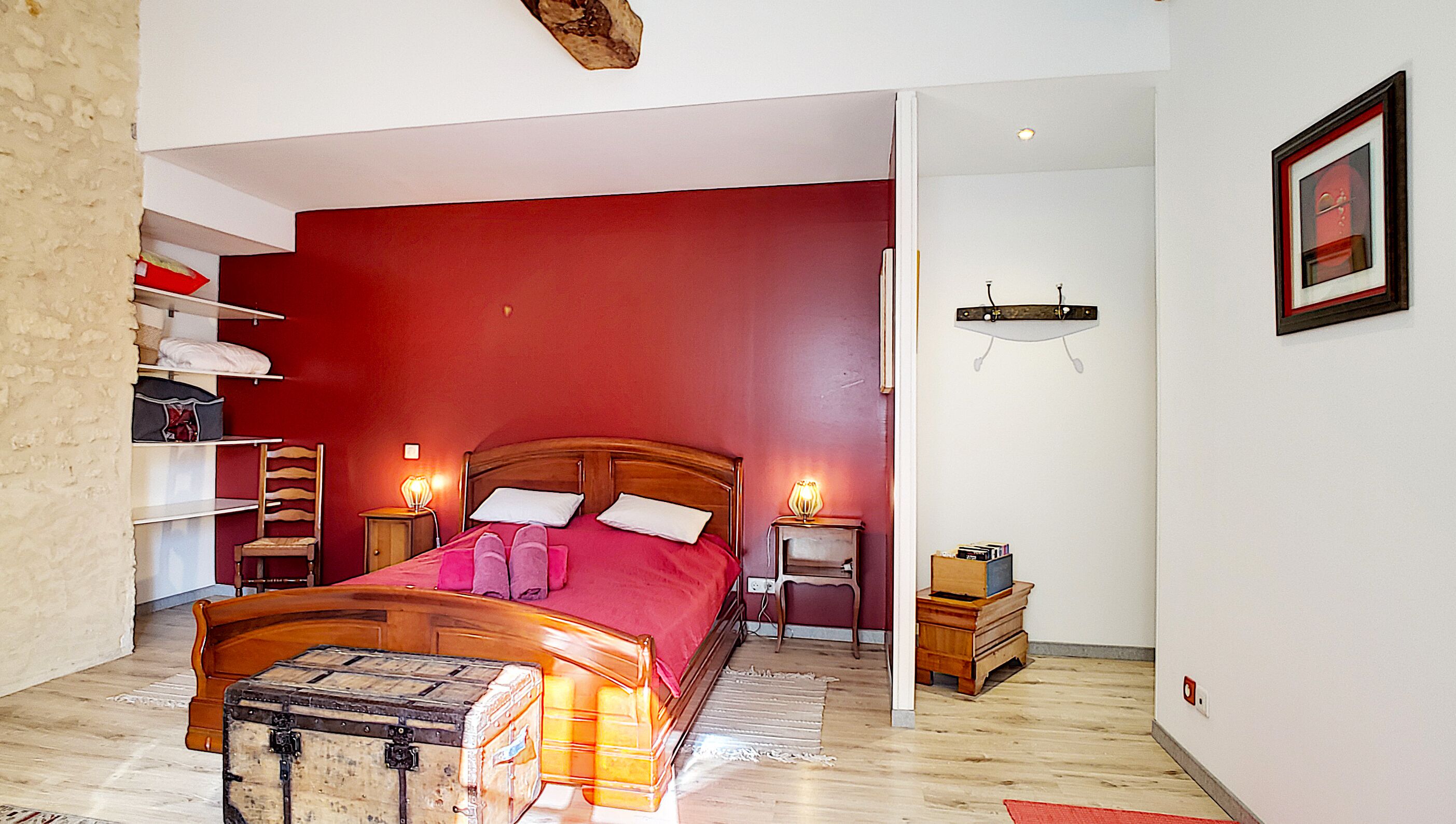 Quarto com cama de madeira, paredes vermelhas, mesa de cabeceira de madeira, luminária, um pequeno armário de madeira e uma mala no chão.