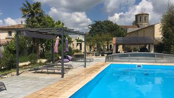 Una piscina con una pérgola y sillones de descanso, rodeada por un jardín y un edificio con una torre de campanas.