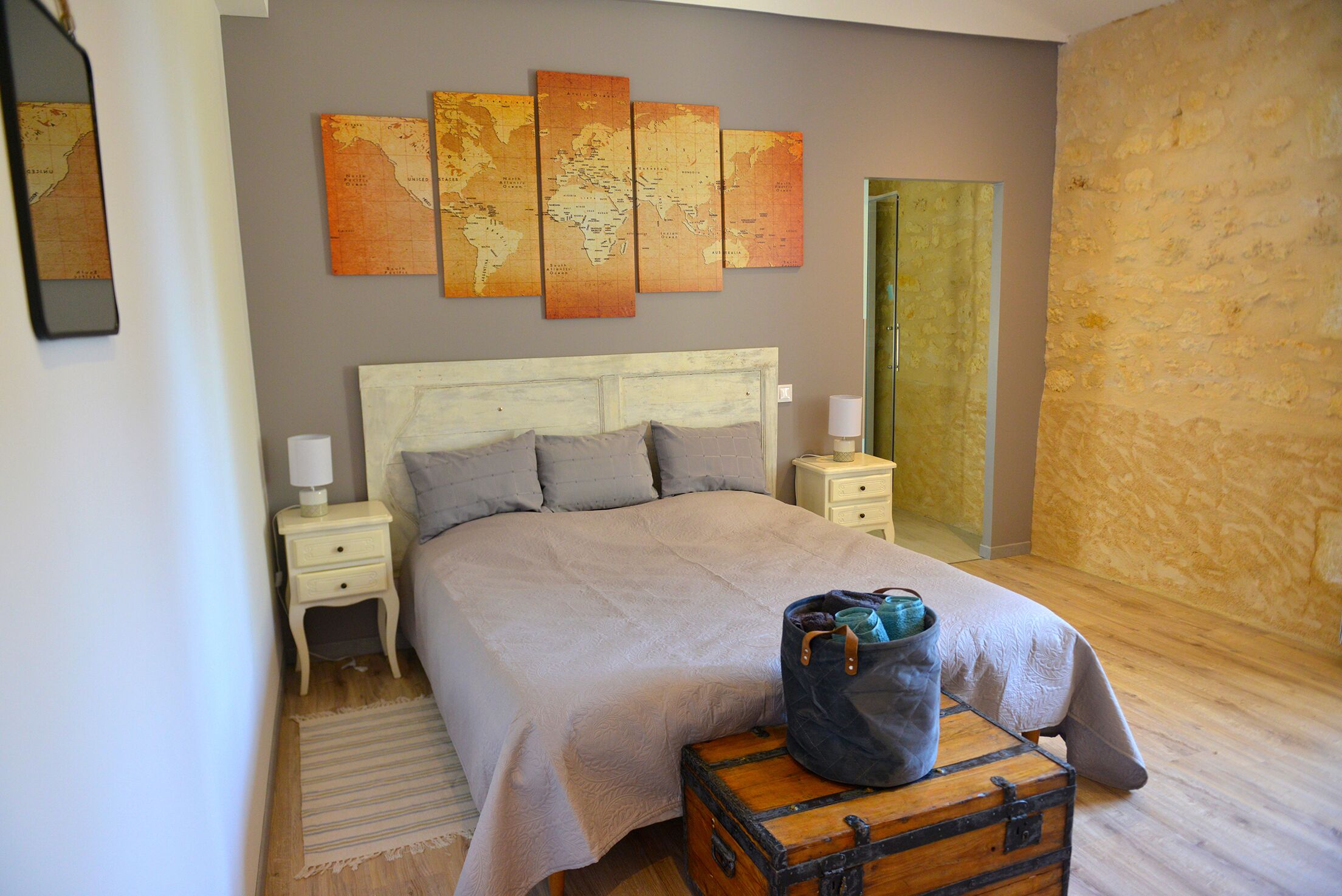 A neatly made bed with a light-colored headboard, a world map on the wall, and a suitcase on the floor.