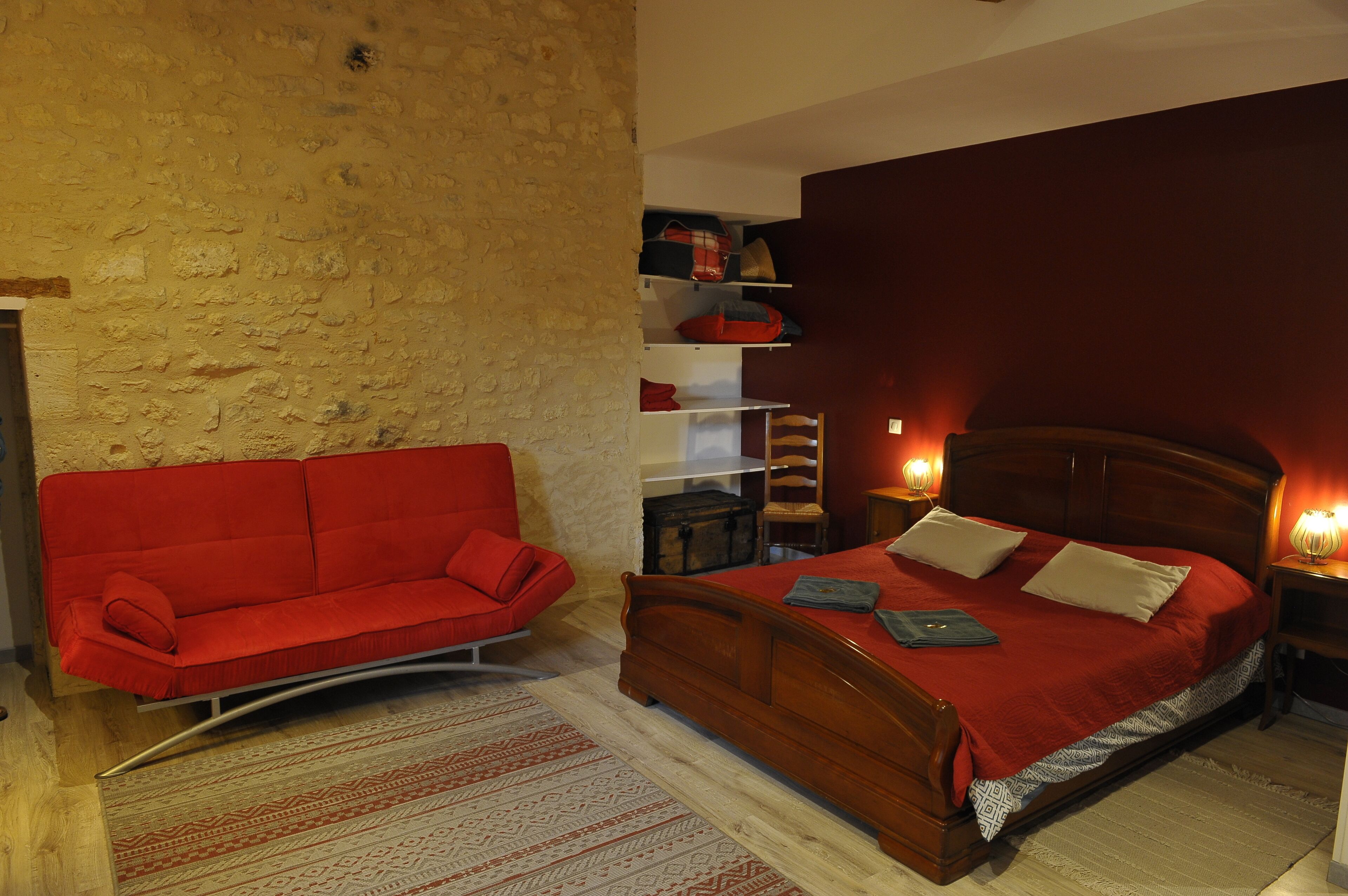 Un dormitorio con una cama de madera, un sofá rojo, un mueble pequeño de madera y una pared de ladrillo.