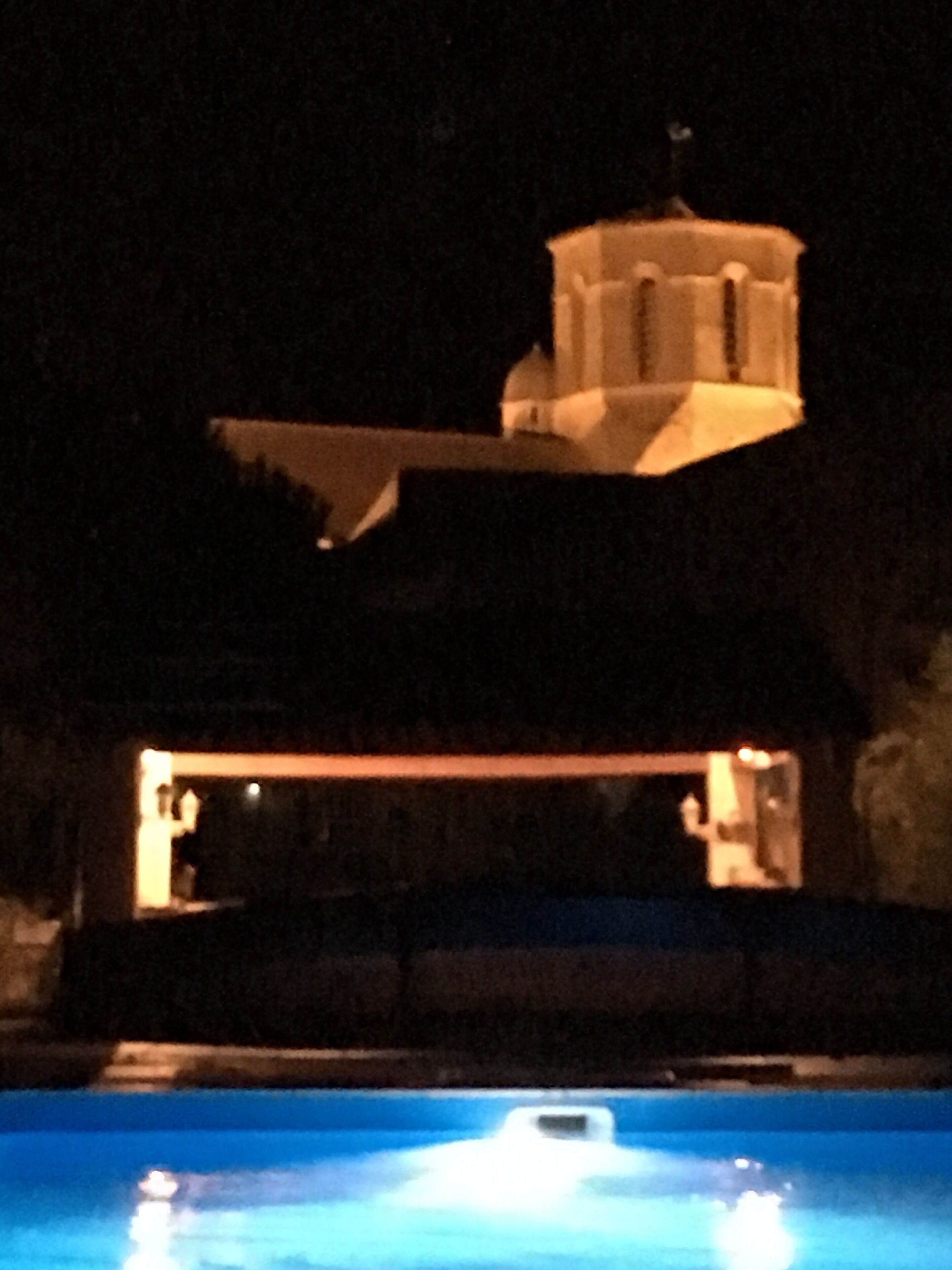 Vista nocturna de un edificio con una estructura tipo torre en la parte superior, una piscina en primer plano y un área iluminada que parece ser recepción o vestíbulo.