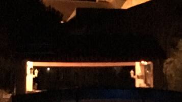 Vista nocturna de un edificio con una estructura tipo torre en la parte superior, una piscina en primer plano y un área iluminada que parece ser recepción o vestíbulo.