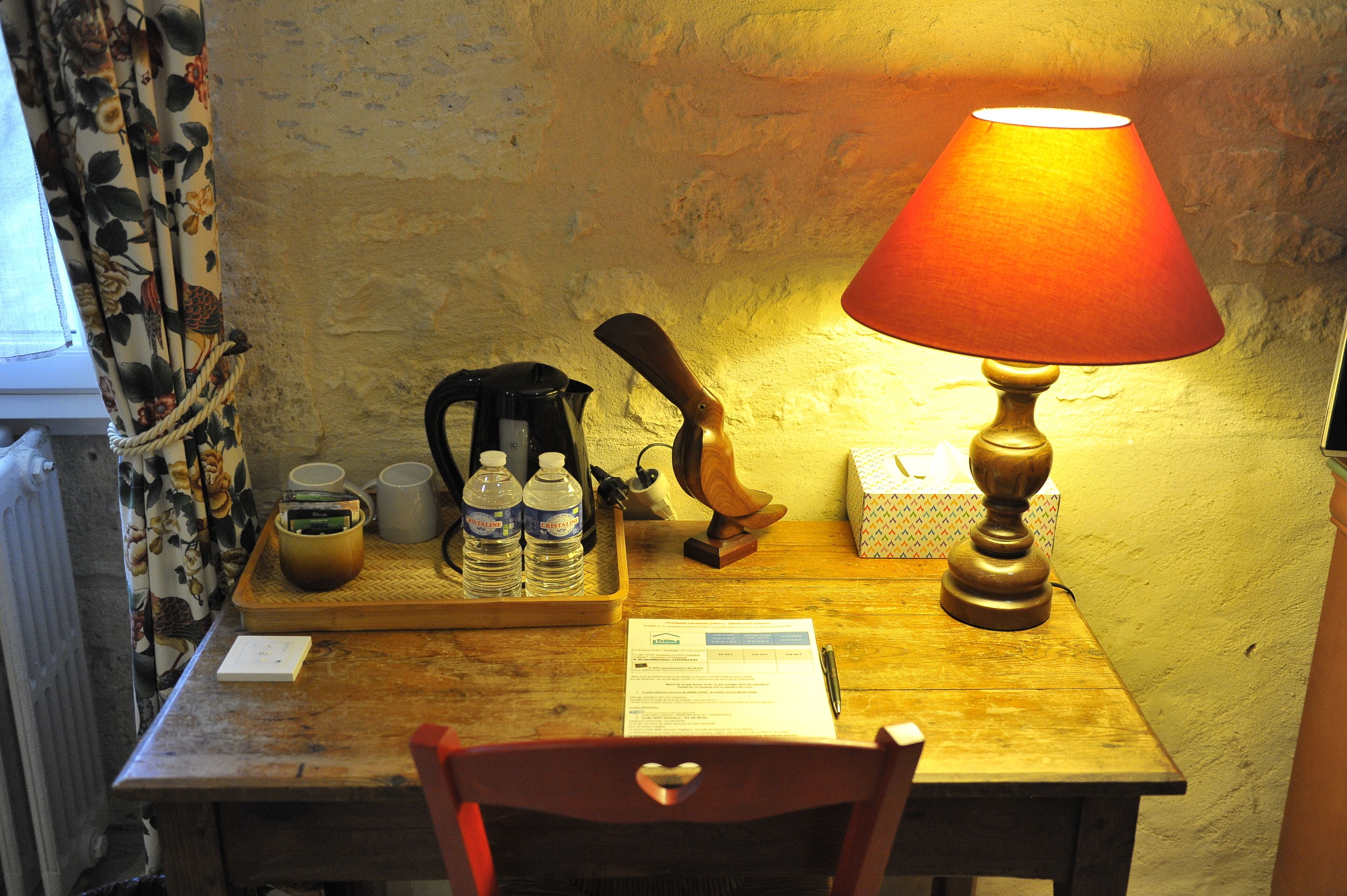 Une table en bois, une lampe, une bouilloire, une machine à café, deux bouteilles d’eau, une tasse, une boîte et une chaise.