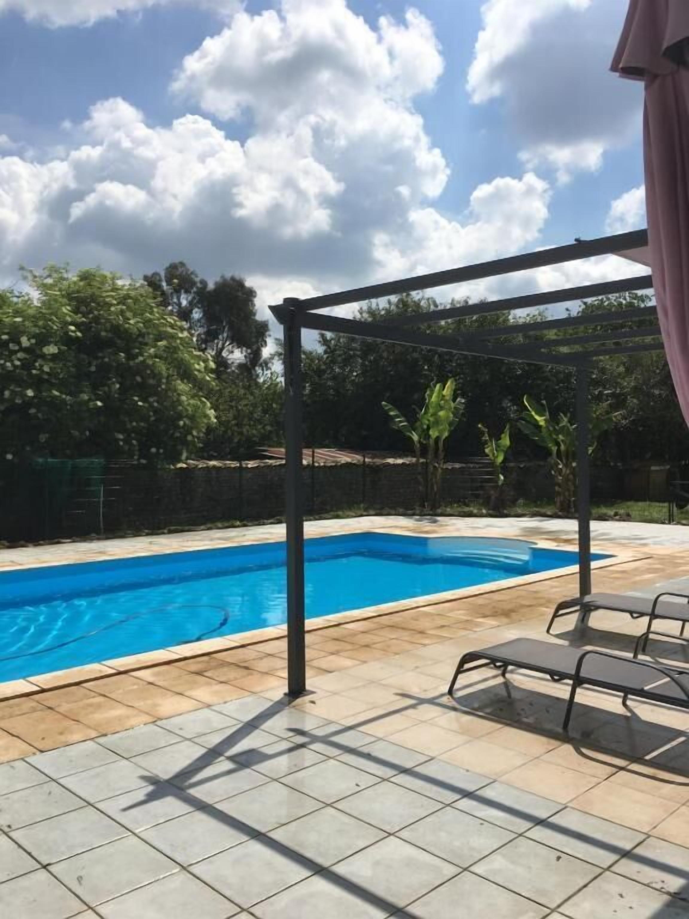 En swimmingpool med en pergola och solstolar, omgiven av en plattlagd uteplats och grönska.