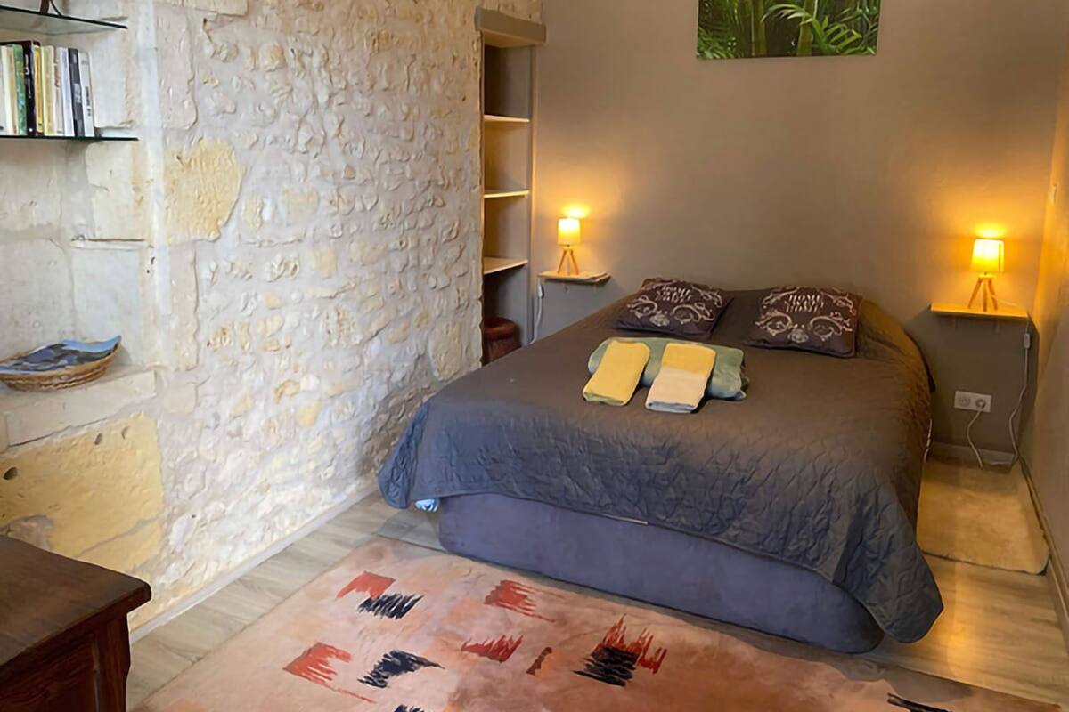 A bedroom with a stone wall, a bed with a blue bedspread, two bedside lamps, and a bookshelf with books.