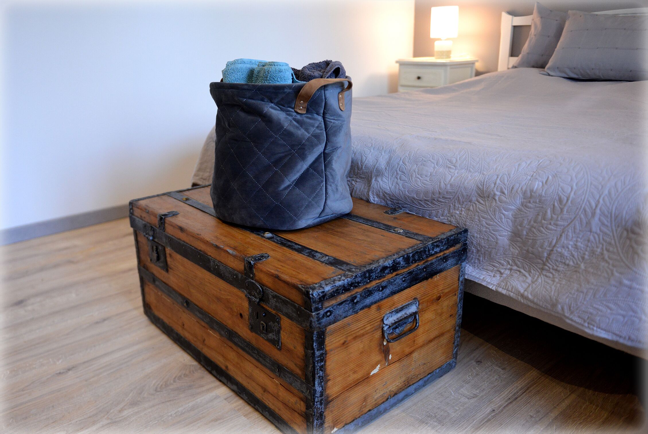 A wooden trunk with a quilted bag on top, placed on a wooden floor in a bedroom with a bed and nightstand in the background.