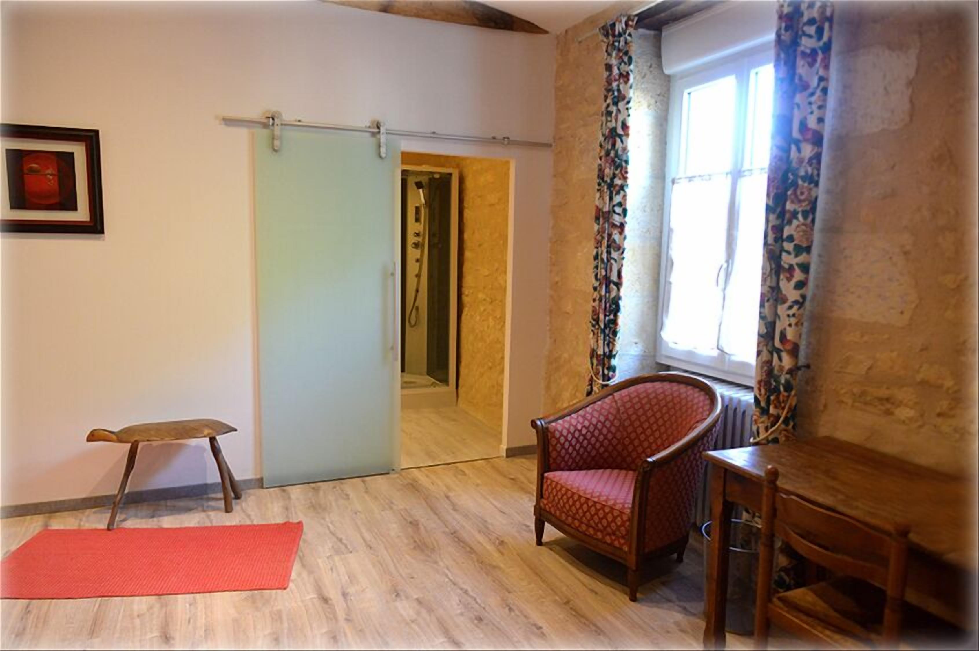 A room with a wooden floor, a red chair, a wooden table, a red mat, a door, and a window with curtains.
