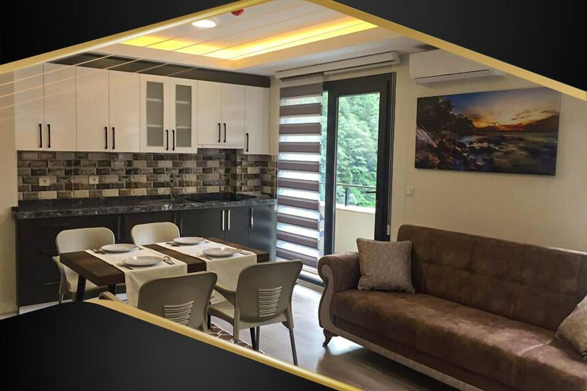 Una cocina moderna con armarios blancos, encimera negra y un comedor con mesa y sillas. Hay un sofá marrón y un cuadro en la pared.