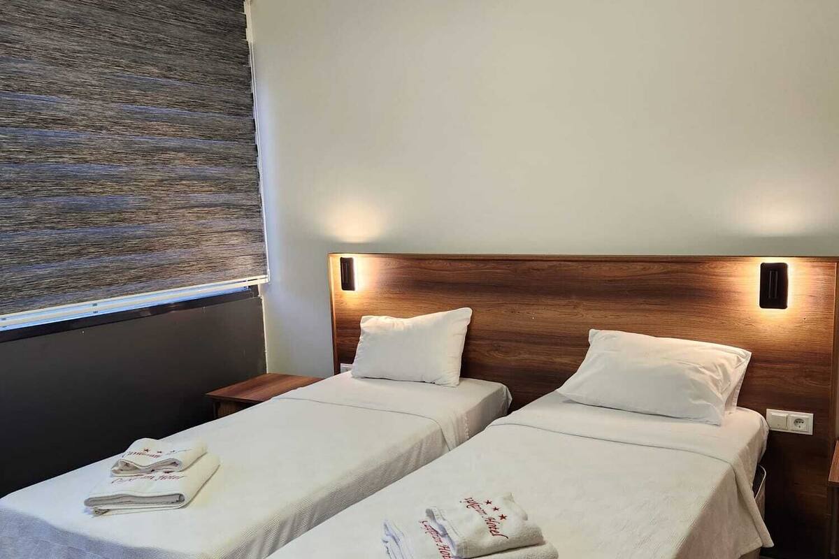 Habitación de hotel con dos camas, cada una con una toalla doblada y un almohadón. La habitación tiene un diseño moderno con cabecera de madera y una lámpara fijada a la pared.