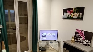 A living room with a television displaying 'LOUANE SENTIMENTS' and 'MAINENTANT DISPONIBLE'. There is a glass cabinet with a remote control, a small statue, and a picture frame with flowers. - Location Saisoniere au Calme Dans Petite Copropriete (Publier)