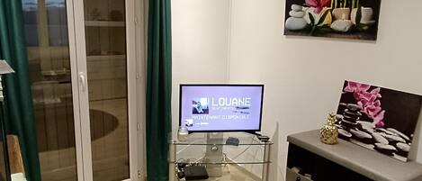 A living room with a television displaying 'LOUANE SENTIMENTS' and 'MAINENTANT DISPONIBLE'. There is a glass cabinet with a remote control, a small statue, and a picture frame with flowers.