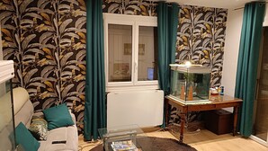 A living room with a sofa, a coffee table with magazines, a fish tank, and a window with curtains. - Location Saisoniere au Calme Dans Petite Copropriete (Publier)