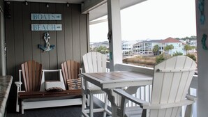 Ett avskärmat utomhusområde med sittplatser, Adirondack-stolar och ett bord, skyltar som visar vägen till "BÅTAR" och "STRAND", och utsikt över ett bostadsområde med hus och en vattenyta.
