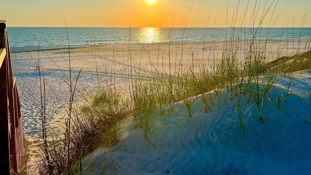 Ein Strand mit untergehender Sonne über dem Meer, mit Sanddünen und spärlicher Vegetation.