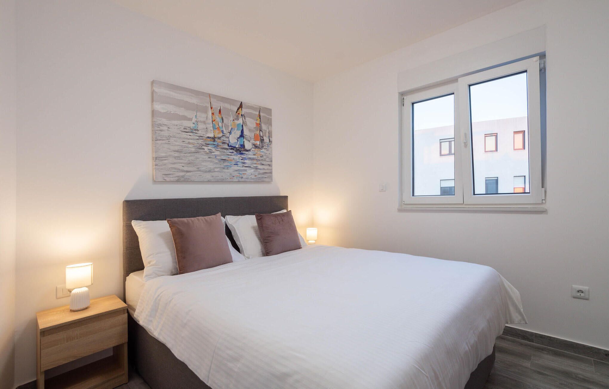 Ein Schlafzimmer mit einem Bett, zwei Nachttischen, einer Nachttischlampe, einem Fenster mit Blick auf Gebäude und einem Bild an der Wand.