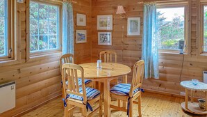 Una cabaña de madera con una mesa redonda también de madera y cuatro sillas con cojines a rayas. Hay cuadros en las paredes y una mesita con una lámpara.