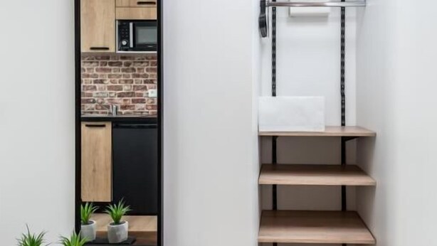 Een smalle gang met een houten tafel, potplanten en een paar schoenen. Een spiegel weerspiegelt een keuken met een bakstenen muur en moderne apparaten.