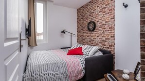 Een slaapkamer met een bakstenen muur, een bed met beddengoed met een patroon, een zwarte stoel, een nachtkastje met een lamp en een klok aan de muur.