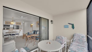 Ein Zimmer mit rundem Tisch, Stühlen, einer Sitzecke und einer Küchenzeile, verbunden durch eine Glastür, die in einen weiteren Raum führt.