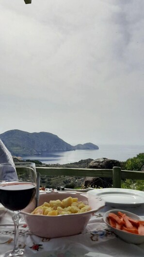A table with a glass of red wine, a bowl of potatoes, and a plate of carrots, overlooking a mountainous landscape and the sea.
