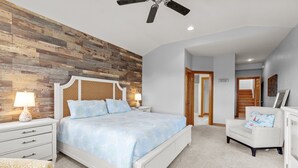 A bedroom with a wooden accent wall, a bed with a blue bedspread, a white nightstand with a lamp, a white armchair, and a ceiling fan. - 1603 - Bare Feet Retreat - Pet-Friendly Corolla Retreat Near the Beach (Corolla)