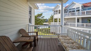 A wooden deck with two wooden chairs and a cushioned bench, overlooking a lake and a white building with a balcony. - 1603 - Bare Feet Retreat - Pet-Friendly Corolla Retreat Near the Beach (Corolla)