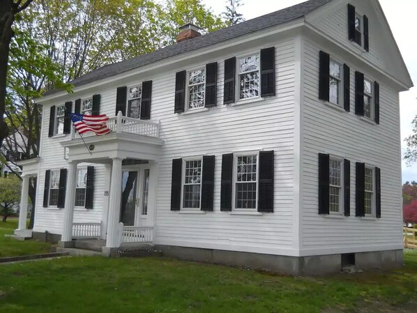 Uma casa branca de dois andares com persianas pretas, varanda e uma bandeira americana.