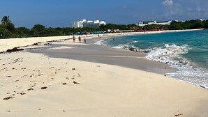 Una playa con agua azul cristalina, orilla arenosa y algunas personas caminando.