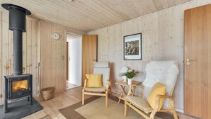 Una habitación acogedora con una estufa de leña, dos sillas de madera con cojines amarillos, una mesita con un jarrón y un cuadro en la pared.