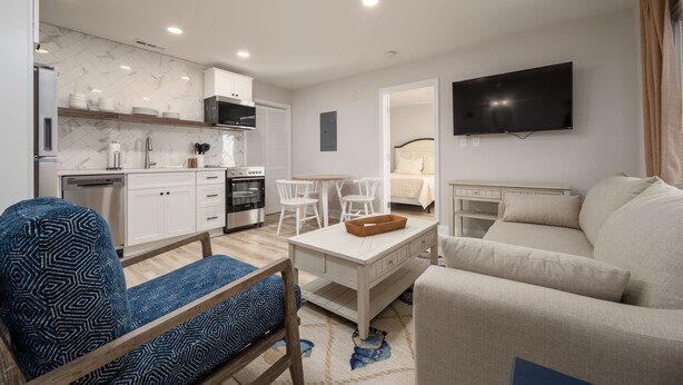 A modern living room with a sofa, a dining table, and a kitchen area with white cabinets and a flat-screen TV.