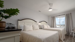A hotel room with two beds, a ceiling fan, a large window with curtains, and a potted plant.