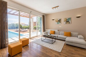 Un vaste salon doté d’une grande porte-fenêtre coulissante donnant sur une piscine, d’un canapé blanc agrémenté de coussins orange et d’une table basse.