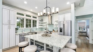 Une cuisine moderne avec des meubles blancs, un îlot central en granit doté de tabourets, des appareils en acier inoxydable et un lustre.