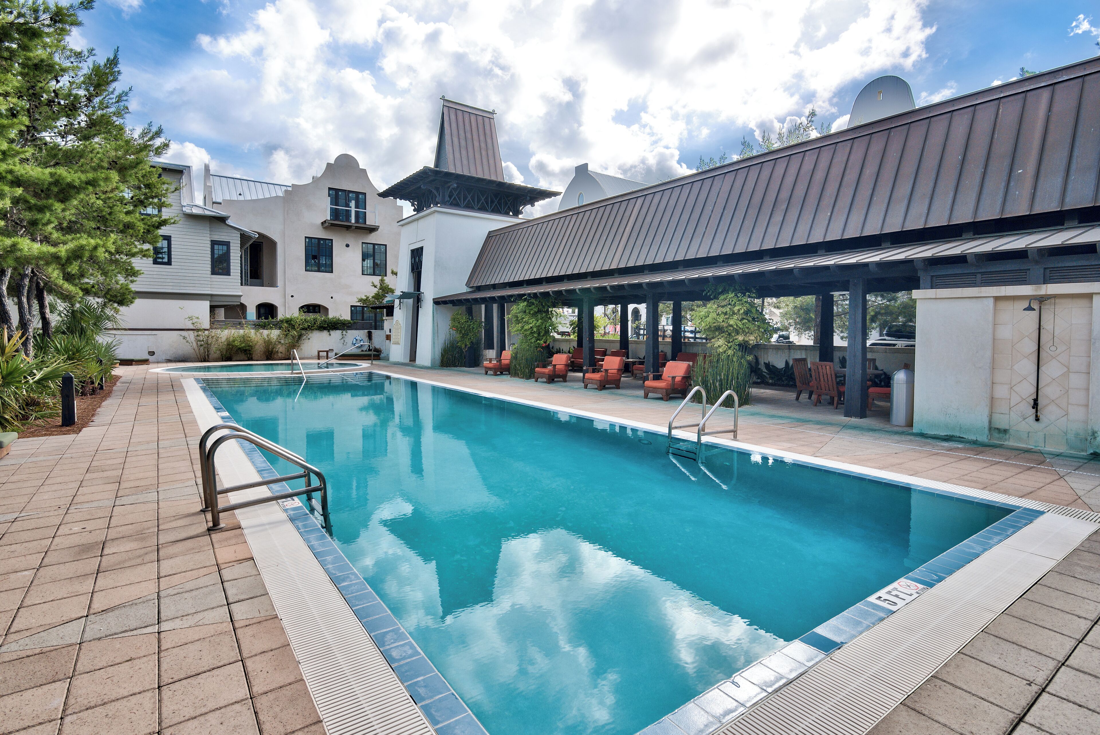 Una piscina con solárium de baldosas, rodeada de mobiliario exterior y un edificio de estilo arquitectónico distintivo.