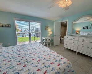 A bedroom with a bed, a dresser, a desk, and a chair. There is a window with blinds, a door, and a view of a garden.