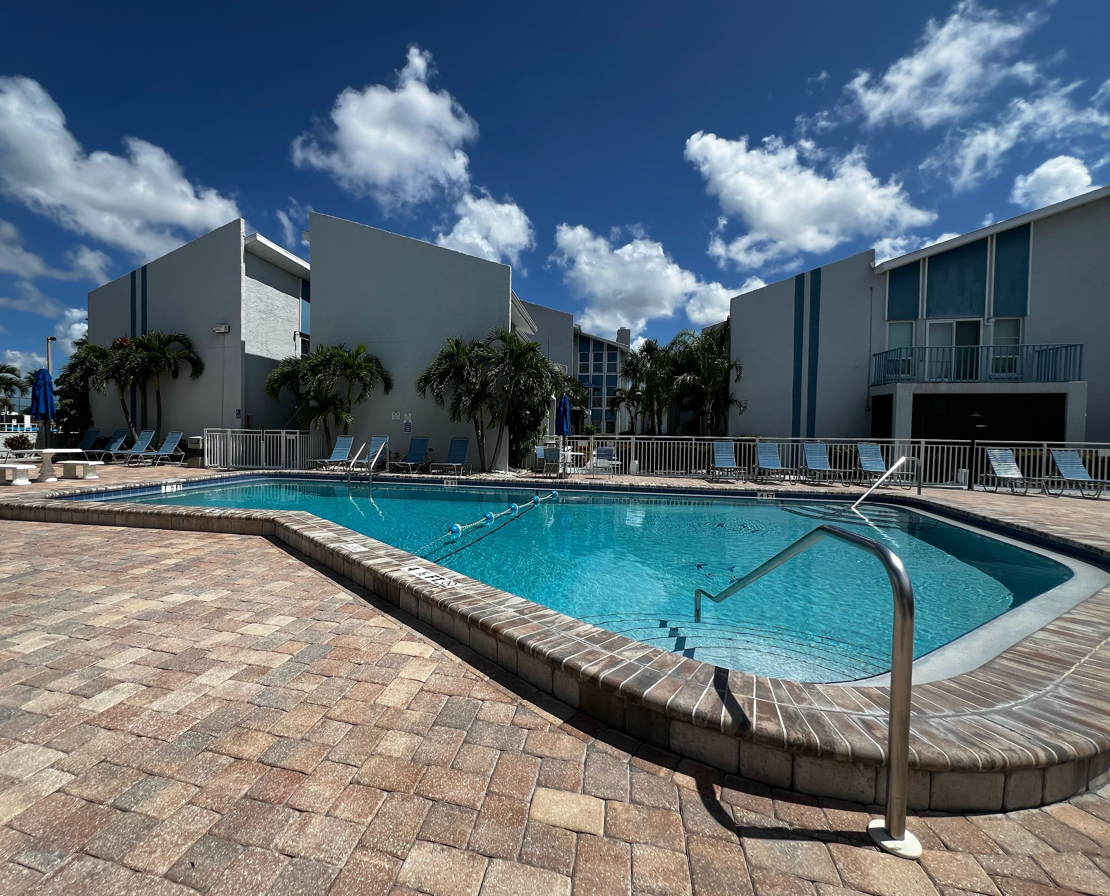A swimming pool with lounge chairs and palm trees, surrounded by buildings with balconies.